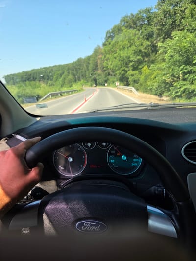 The image, taken from the driver's perspective inside a Ford vehicle, shows a person's left hand, holding a lit cigarette, resting on the steering wheel. The steering wheel features the Ford logo. The dashboard displays various indicators, including a speedometer reading approximately 60 km/h, an odometer showing 347643 km, and a temperature gauge reading 31°C. The road ahead, curving to the left, is bordered by a metal guardrail and orange traffic cones. Lush green trees line both sides of the road, suggesting a rural or suburban setting. The sky is clear and blue. The car is driving on a paved road. The overall impression is of a person driving on a sunny day. The location is described as the city of Venets, Bulgaria.