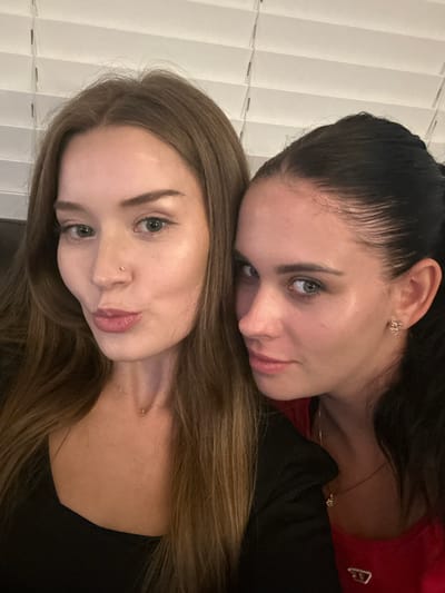 Two young adult females are depicted in a close-up, chest-up portrait, captured indoors in 마포구, South Korea. The individual on the left has long, light brown hair, light-colored eyes, and a small stud piercing in her left nostril. She is wearing a dark top and a thin necklace with small charms, and is observed with her lips slightly puckered. The individual on the right has dark hair pulled back, light-colored eyes, and is wearing a red top under a dark outer garment. A small, light-colored stud earring is visible in her right ear. She is positioned slightly behind and to the right of the first individual, looking directly at the camera with a neutral expression. The background features white horizontal window blinds.