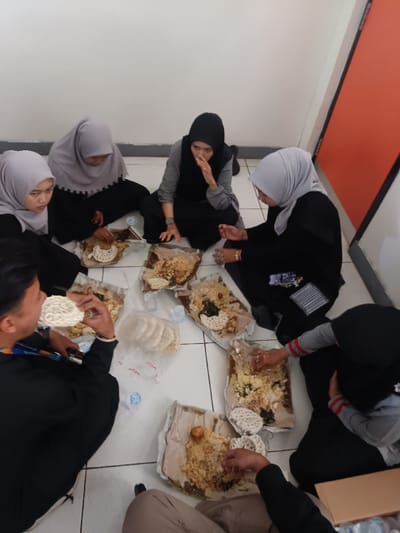 A group of seven people are sitting on a tiled floor, arranged in a semi-circle, sharing a meal. Each person has a portion of food spread out on a paper wrapper in front of them, with what appears to be rice, vegetables, and crackers on top. Several of the individuals are wearing head coverings, and they seem to be enjoying their meal together in a casual, indoor setting.