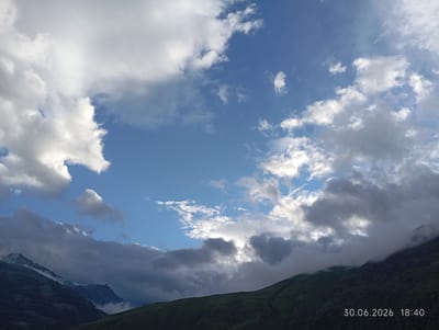 This is a landscape photograph taken in Tyrnyauz, Russia. The scene depicts a dramatic sky filled with a mix of white and dark grey clouds, with patches of bright blue sky visible. The clouds create a dynamic and somewhat moody atmosphere, with some areas brightly lit and others casting shadows.
In the foreground, rolling green hills dominate the lower portion of the image. These hills are covered in lush vegetation, with some areas appearing darker, suggesting shadows or denser foliage. In the background, majestic mountain peaks rise up, some partially shrouded in mist or clouds. The mountains have a rugged, imposing appearance.
The time of day appears to be late afternoon or early evening, as indicated by the lighting conditions and the timestamp in the lower right corner. The weather seems to be variable, with the presence of both sunshine breaking through the clouds and darker, more ominous clouds suggesting a chance of rain or a change in conditions. The overall mood is one of natural grandeur and a sense of the wildness of the mountainous landscape.
Visible text in the lower right corner reads: "30.06.2026 18:40". This indicates the date and time the photograph was taken. There are no people or man-made objects prominently featured in the image, focusing purely on the natural scenery.