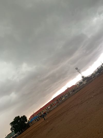 The image depicts an outdoor scene, likely a schoolyard or open field, under a heavily overcast sky. The ground is a reddish-brown dirt or sand, with some scattered debris. In the background, a long, multi-story building with an orange-tiled roof runs horizontally across the frame. This building appears to be an educational or administrative facility, with visible windows and some signage. To the left of this main building, a smaller structure with a blue roof is present, and a large, leafy green tree stands nearby. A tall telecommunications tower with multiple antennas is visible to the right of the orange-roofed building. A few individuals are discernible in the middle ground, some appearing to be standing or walking. The overall impression is of a slightly overcast day, possibly before or after rain, with a utilitarian urban or semi-urban setting. The image is tilted, with the horizon running diagonally from the lower left to the upper right. The specific location of Kagwaru, Nigeria, is not visually evident from the image itself, as there are no distinct landmarks or signage to confirm this detail.