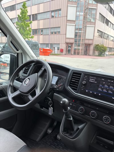 This image provides an interior view of a Volkswagen van, likely a Crafter, in Mannheim, Germany. The perspective is from the driver's seat, looking towards the dashboard and out the windshield. The dashboard features a steering wheel with the Volkswagen logo and airbag indicator, a gear shift lever, climate control knobs, and a touchscreen infotainment system displaying the date "30.06.2026", time "8:18", and "Willkommen". Outside the vehicle, a modern commercial building with a glass elevator shaft and pink-tinted facade is visible. In front of the building, an orange dumpster and bicycles parked along a sidewalk suggest an urban or industrial setting. The overall scene indicates a daytime snapshot of the vehicle's interior and its immediate surroundings.