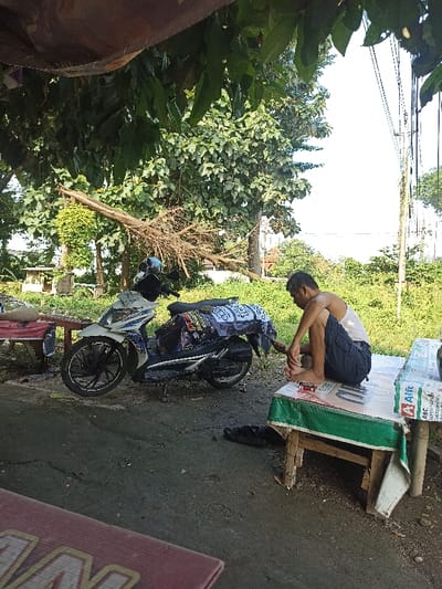 The scene takes place in a residential area in Kecamatan Kartasura, Indonesia, with coordinates of -7.56 latitude and 110.763 longitude. A man in a white tank top and black shorts is sitting on a wooden bench with a green and white tablecloth. He is looking down at his feet and appears to be wearing sandals. A white motorbike is parked behind him with its rear tire facing him. The motorbike has a black and blue seat cover and black handlebars. The man appears to be sitting on the bench to the right of the motorbike. There is a brown wooden table in the foreground, which is likely a coffee table or a small table. The background is a lush green, and there are several trees in the background, and a brown awning in the left foreground. The scene is likely a typical residential neighborhood. The man's actions and the presence of the motorbike and table suggest that the scene is likely a casual setting, such as a backyard or a porch. The man's relaxed posture and the presence of a tree and the awning suggest a comfortable and peaceful atmosphere.