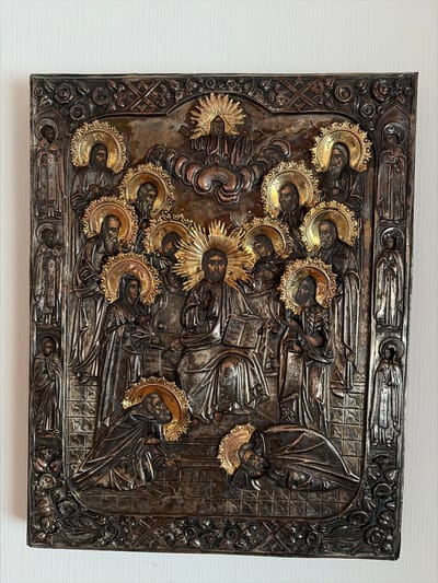 This is a photograph of a silver icon depicting Jesus Christ enthroned, surrounded by apostles and saints. Jesus, in the center, is shown with a golden halo and is holding an open book. The apostles and saints are also depicted with halos, some standing and others kneeling. The icon is intricately detailed, with ornate borders featuring floral motifs. The metallic surface of the icon shows signs of age and wear, with areas of patina and tarnish. The background is a plain white wall, which serves to highlight the icon itself. The image is taken from a straight-on perspective, with the icon filling most of the frame. There is no discernible location in the city of Osnabrück, Germany, visible in this image.