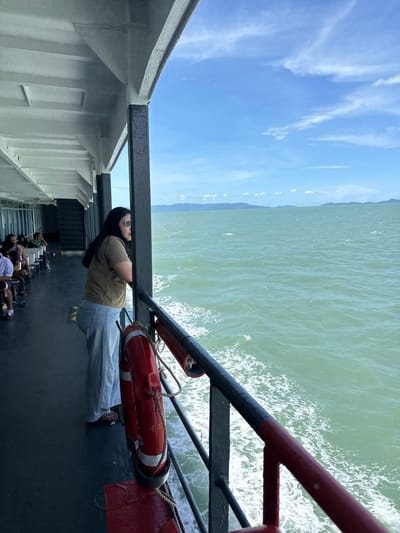 In Thong Nian, Thailand, a woman stands on the deck of a ferry, gazing out at the calm, turquoise waters. The ferry deck is dark gray, with white overhead structures and black support beams. A red and white life preserver hangs on the railing. In the distance, the hazy outlines of islands are visible against a bright blue sky dotted with wispy clouds. Several other passengers are seated on benches further down the deck, some blurred due to the ferry's motion. The woman is wearing a brown t-shirt and light blue jeans, with sunglasses on her head. The water churns with white foam from the ferry's wake.