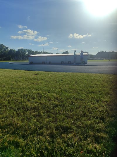 The image depicts a bright, sunny day at a sports facility, likely a running track. Two young individuals, a boy in a green shirt and shorts and another in a blue shirt and shorts, are standing on top of a long, white, rectangular structure with a red roof. The structure appears to be a changing room or storage facility. One boy is looking at the other, who is holding a water bottle and looking away. The background shows a blue running track, a grassy field, trees, and buildings in the distance. The sun is high in the sky, casting a bright glare. The overall setting suggests a casual, outdoor activity in Tczew, Poland.
