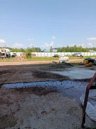 The image depicts an outdoor industrial or construction area in Borodino, Russia. In the foreground, a dirt and concrete ground surface is visible with puddles of water and scattered debris. Further back, there is a pile of gravel and a yellow compaction machine covered with a tarp. Several vehicles, including a truck and what appears to be a small utility vehicle, are parked in the mid-ground. A white fence runs along the edge of the property, with a line of trees and industrial structures visible beyond it. The sky is clear with some scattered clouds. The overall scene suggests a working environment with building materials and equipment.