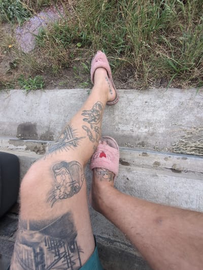 The image shows a close-up view of two tattooed legs, wearing pink fuzzy slippers with red hearts. The legs are positioned on a concrete ledge next to a patch of overgrown grass and dirt. The tattoos are visible on both legs, with various designs including what appears to be a building or bridge structure, a feline face, and abstract patterns. One slipper is on the left leg, near the ankle, and the other is on the right leg, further down the leg. The scene appears to be outdoors, possibly in a residential or rural area given the presence of grass and uneven ground. There are no other people or objects clearly identifiable in the frame. The lighting suggests it is daytime. The context of Perliavka, Ukraine is not visually evident from the image itself.