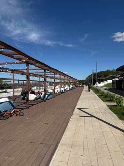 This image captures a vibrant, sunny outdoor scene along a waterfront promenade in Taman, Russia. The perspective looks down a long, modern leisure area.

In the foreground and extending into the mid-ground, a wide wooden deck runs parallel to the visible body of water. This deck is covered by an open pergola-like structure made of dark brown wooden beams, from which string lights are suspended, suggesting it's also used in the evenings. Numerous light blue and white outdoor lounge chairs or beanbag seats are arranged along this deck, providing ample seating. Several people are visible sitting on these chairs, appearing relaxed and enjoying the sunny weather. In the foreground, an orange bicycle lies on its side on the wooden deck.

Adjacent to the wooden deck, a paved pathway made of light-colored, rectangular tiles stretches into the distance. This pathway is lined with several tall, slender, dark lampposts or decorative poles. Further beyond the pathway, there's a strip of green grass and bushes, with more substantial greenery and hints of buildings in the background. A person dressed in dark clothing is visible walking or standing on the paved path in the mid-distance.

The sky is a clear, brilliant blue with a few scattered, wispy white clouds. Bright sunlight casts strong, distinct shadows from the lampposts and structures, indicating it is daytime, likely mid-morning or afternoon. The body of water, which appears to be a sea or large lake, is visible beyond the wooden deck, reflecting the bright sky. The overall impression is one of leisure and relaxation in an inviting outdoor setting. No discernible text is visible within the image.