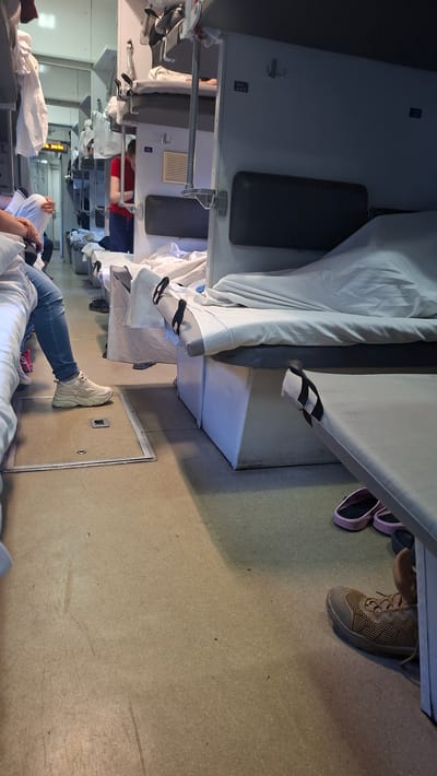 The image shows the interior of a train compartment with several people present. The compartment appears to be a sleeping car, with bunk beds made up with white linens. One person is seated on the lower bunk, wearing jeans and white sneakers, with their legs extended. Another person's feet are visible in black and white striped slide sandals. Further down the aisle, a person in a red shirt and blue shorts is lying on a bunk. In the foreground, a pair of pink slides are visible. A pair of brown lace-up shoes are also on the floor. The scene suggests a journey in progress. The lighting appears to be artificial, indicating either nighttime or an interior setting with limited natural light. There is a label visible on the wall with "22A" and another with "18c." The overall impression is one of a functional and somewhat cramped travel environment. There are no discernible weather cues or explicit indications of time of day beyond the artificial lighting. The context provided indicates the location is Serbynivtsi, Ukraine.