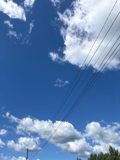 This media file captures a bright, sunny day in Kuvshinovo, Russia, as indicated by the clear blue sky and fluffy white clouds. The perspective is looking upwards, emphasizing the expanse of the sky. Multiple dark utility wires stretch diagonally across the frame from the left to the right, adding a graphic element to the natural scene. In the lower right portion of the image, a utility pole with a light fixture is visible, further confirming the setting as an outdoor, likely semi-urban or rural, environment. The sun is high in the sky, suggesting it is midday. There are no people or animals visible in the frame, and no text is present. The overall impression is one of a serene and ordinary day.