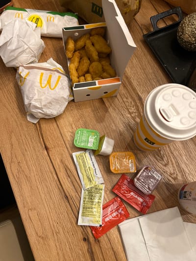 The scene depicts a fast-food meal from McDonald's spread across a light brown wooden table surface. In the upper left quadrant, several items wrapped in white paper with yellow McDonald's 'M' logos are present, including one partially visible burger or sandwich. Adjacent to these, an open white cardboard box with subtle yellow and dark-striped patterns along the bottom edge contains numerous golden-brown chicken nuggets. In the upper right, a black textured rectangular tray with handles holds a round, textured, golden decorative object. Towards the right, a white and yellow striped McDonald's cup with a white lid is positioned upright, containing a beverage. Numerous condiment packets and small containers are scattered across the table, including green-capped "Creamy Cheese" sauces (one upright, one overturned), yellow "Pommes-Frites Sauce" packets (with French text), an orange-capped "Sauce Classic McDonald's" (Moutarde Mayonnaise), a dark-capped sauce, and multiple red "Tomato Ketchup" packets. White paper napkins are visible in the lower right. A brown McDonald's paper bag is partially visible in the upper background, and a light-colored upholstered chair back is partially visible in the lower left. The presence of McDonald's branding and French text on some condiment packets indicates the meal was purchased from a McDonald's restaurant in France. Given the context provided, the items are located within the city of Corbeil-Essonnes, France, likely in a private indoor setting, as no external city landmarks are discernible.