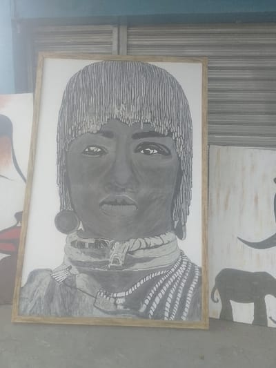 A large, vertically oriented, wooden-framed art canvas depicting a monochrome portrait is centrally positioned on a grey concrete surface, indicative of a pavement or floor, in Bamburi ward, Kenya. The portrait, rendered in shades of grey, black, and white, features an individual with a distinctive fringe-like hairstyle covering the forehead, large circular earrings, and multiple decorative necklaces or beaded strands around the neck and upper chest. To the immediate left, a partial view of another canvas displays reddish-brown and dark elements. To the right, a third canvas with a distressed white background presents dark grey silhouettes, including an elephant in the lower portion and an abstract shape above it. The immediate background behind the canvases consists of a closed, grey metallic rolling shutter, topped by a section of a blue-painted wall or overhead structure. No individuals are present; the canvases are stationary, displayed upright.