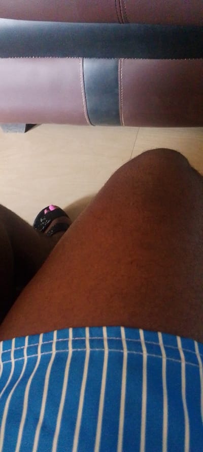 This media file shows a close-up view of a person's legs and thighs, wearing blue and white striped shorts. The person is also wearing black sandals with pink accents on their toes. In the background, a portion of a brown and black upholstered couch is visible, suggesting an indoor setting. The lighting appears to be natural, possibly from a window, casting subtle shadows. The image is taken from a low angle, focusing on the lower half of the body. The scene likely takes place during the daytime. No text is visible in the media file. The overall context suggests a casual, everyday moment in a home environment in Gboko, Nigeria.