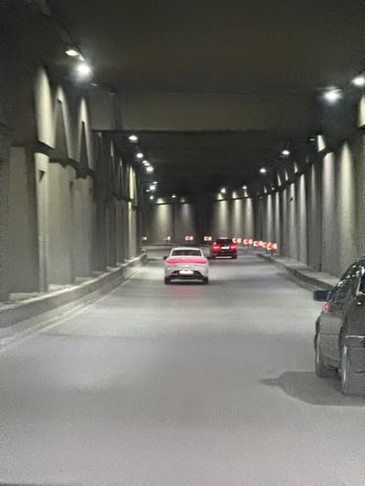 The image shows a view from inside a concrete tunnel in Baku, Azerbaijan. The tunnel is illuminated by a series of lights mounted on the ceiling. The walls of the tunnel are lined with concrete pillars. Several cars are visible on the road within the tunnel. In the foreground, the rear of a dark-colored car is on the right side. Further ahead, a white car is in the middle lane, and behind it, a red car is in the left lane. Warning signs indicating a curve to the left are placed along the right side of the road. The road surface appears to be asphalt and is slightly wet, reflecting the tunnel lights. The overall atmosphere is dark and somewhat enclosed due to the tunnel structure.