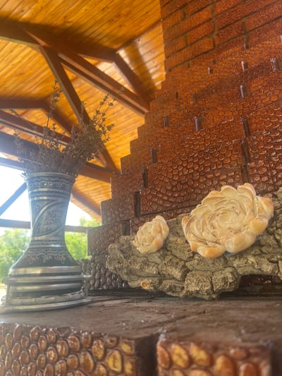 The image captures a close-up of an outdoor area with a decorative brick structure and a wooden ceiling. A tall, ornate vase filled with dried lavender stands to the left. The brickwork is a rich brown, with each brick featuring a pattern of raised, rounded shapes. Two shell decorations, resembling flowers, are placed on a piece of rough wood attached to the brick structure. The wooden ceiling, with visible beams, is illuminated by natural light, suggesting it's daytime. In the background, blurred greenery indicates an outdoor setting, possibly a garden or natural landscape. The scene is devoid of people. The location is identified as Bolshoy Molokish, Moldova, though the specific context within the town is not discernible from the image.