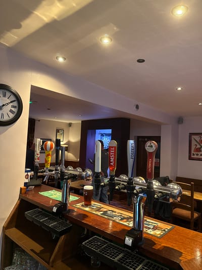 This image depicts the interior of a pub, identified as being located in Sigglesthorne, United Kingdom.

The foreground features a dark, polished wooden bar counter. Numerous chrome beer taps are mounted along the bar; prominent handles include Cruzcampo, Amstel, Fosters, and L'Autentica (Birra Moretti), with other less distinct handles, including one with a red apple design and another yellow/orange striped, on the left. A pint glass containing a dark amber liquid (beer) rests on a branded bar mat to the right of the central taps. Below the taps, two black grates serve as drip trays. A large, black-framed wall clock with Roman numerals is affixed to the pale wall on the far left, displaying approximately 12:45. The ceiling is white with multiple recessed downlights providing illumination.

Beyond the bar, the pub interior extends into an area with wooden seating, including benches and tables. In a recessed alcove or doorway at the rear, a male individual is partially visible from the back, wearing a dark top and light-colored trousers, standing in front of a dartboard mounted on a wall. To the far right, a framed picture featuring two red swans on a white background hangs on a pale wall above a wooden bench. The overall environment is illuminated by warm lighting from the ceiling fixtures.