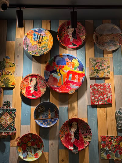 The image captures a wall in what appears to be a decorative setting in Tsakhkadzor, Armenia. The wall is made of alternating horizontal wooden planks in light brown and blue-grey shades. A diverse collection of decorative ceramic plates and tiles are mounted on the wall. The plates have various painted scenes, some of people, and others of landscapes or abstract patterns. One plate even functions as a clock. There are also square ceramic tiles adorned with floral and botanical motifs. The overall arrangement of the art appears to be random. A black cylindrical light fixture is mounted on the wall on the left. A security camera is barely visible in the lower-left corner.