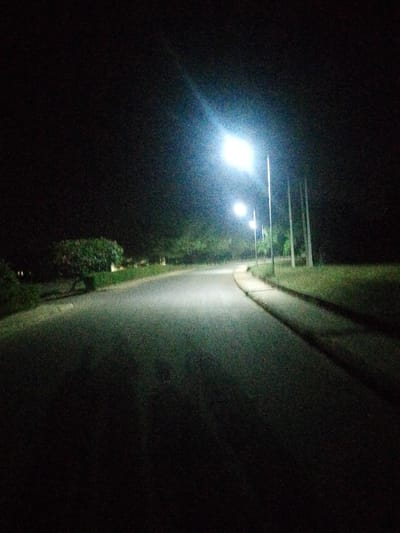 A dark, curving road illuminated by streetlights stretches into the distance. On the left, a tree and shrubbery line the road. A sidewalk borders the right side of the road, with grass beyond it. The scene is dark and somewhat grainy, suggesting nighttime. The nearest streetlight is bright, casting a glare and reflecting on the pavement. The overall impression is of a quiet, suburban street at night.