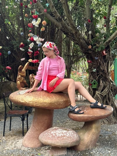 A young woman is sitting on a mushroom sculpture in an outdoor setting. The mushroom sculpture is brown and tan, with a smooth, shiny finish. A smaller, golden rabbit sculpture is positioned near her on the mushroom. The woman is wearing a pink button-up shirt over a pink top, a red skirt with white polka dots, black sandals with white accents, and a patterned headscarf. She has glasses and is looking down. The background features a lush, tree-filled area with hanging decorative flowers. The ground is covered in small rocks. A chair is visible next to the mushroom sculpture. The location is Nam Nha Trang, Vietnam.