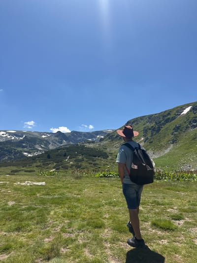 A person wearing a hat, t-shirt, and denim shorts stands in a grassy field, looking out at a mountainous landscape. The person has a backpack on and is facing away from the camera. The mountains in the background are green with patches of snow and rock. The sky is clear blue with a bright sun. The overall scene is a natural, outdoor environment.
