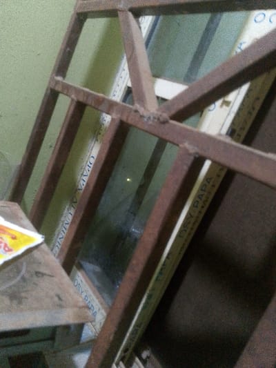An angled view showcases a brown, welded metal frame, partially obscuring a window. The frame is composed of rectangular bars, forming multiple square and rectangular openings. Behind the frame, a partially visible white and yellow window frame is inscribed with "DSY PAPA ALUMINIO". The background features a green wall, slightly blurred and out of focus. The scene is dimly lit, casting subtle shadows and highlighting the textures of the materials.