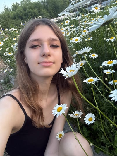 This is a medium shot selfie of a young woman lying down amongst daisies. She has long, light brown hair, fair skin, and is wearing a black tank top. Her eyes are dark brown and she has on eyeliner. Her lips are slightly parted and are a natural pink color. Several daisies are visible, some close to her face and others in the background, with their white petals and yellow centers contrasting against the green foliage and grey ground.
The woman appears relaxed, with a serene expression. The lighting suggests it is daytime, possibly late afternoon given the soft light. The setting appears to be a natural outdoor environment, likely a field or garden, with trees visible in the background. The presence of daisies and foliage indicates a temperate climate. The overall mood is peaceful and idyllic. No visible text is present in the image.