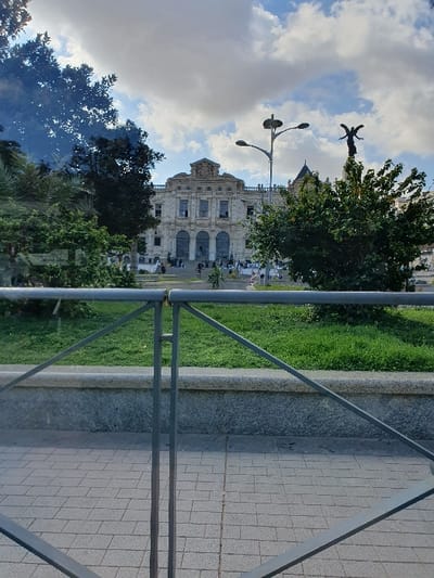 The image shows a view from a low angle looking up through a metal railing toward a building. The building appears to be an older, two-story structure with a light-colored facade and windows. There are trees and plants in the foreground and background. The sky is partly cloudy. The image is taken in Oran, Algeria, at the coordinates 35.704, -0.65. The photo is taken from the sidewalk in front of a building. The railing is in the foreground, and the building is in the background. The building is tall and has a lot of windows. There is a small patch of grass behind the railing. The sky is blue with white clouds.