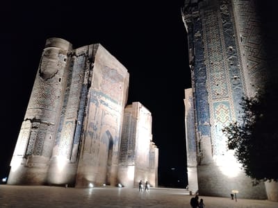 This nighttime photograph captures the imposing facade of the Ak-Saray Palace in Shahrisabz City, Uzbekistan. The massive, ancient structure is illuminated by artificial lights, highlighting its intricate blue and gold mosaic tilework which adorns the towering walls. The architecture is characterized by grand arches and cylindrical turrets, suggesting a historical and possibly defensive purpose. The foreground reveals a paved courtyard area where a few individuals are visible, appearing as small silhouettes against the brightly lit buildings. Two figures are walking in the middle ground, while another group of two, one adult and one child, are closer to the viewer in the lower right corner. The sky above is a deep, dark black, emphasizing the dramatic lighting of the monument. A tree with dark foliage partially obscures the right side of the palace, adding a natural element to the scene. The overall impression is one of awe-inspiring historical grandeur set against the nocturnal darkness.
