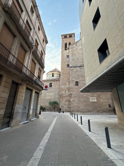An exterior, low-angle, wide shot captures a street scene in Igualada, Spain, featuring a juxtaposition of old and new architecture under a partly cloudy sky. The left side of the image is dominated by the facade of an old building with multiple stories, featuring arched windows with metal railings and roller shutters on the ground floor. A white line demarcates the edge of the brick-paved sidewalk. To the right, a modern building with a beige tiled facade and dark, angular window frames rises up. In the background, a historic stone tower with arched windows and a belfry stands adjacent to a church with a distinctive domed roof adorned with colorful tiles. A small olive tree and a sculpture of a person on a bench are visible in the open plaza area between the buildings. Several dark, angled bollards line the right side of the walkway. The scene is devoid of any visible people.