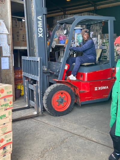 A man in a blue jumpsuit and a brown beanie is operating a red and grey XGMA forklift in what appears to be a warehouse or distribution center. The forklift is laden with what seem to be large rolls of paper or toilet paper, stacked high in the background. The man is looking to his right, slightly away from the camera. A portion of another person wearing a green jacket and a red cap is visible on the right side of the frame, looking towards the forklift operator. In the foreground, stacks of boxes are visible, some with labels indicating "White Cream" and "10 x 1KG". Large orange bottles are also stacked in the background to the left. The environment appears to be an indoor industrial setting with a concrete floor. The scene suggests an active work environment within a commercial or industrial facility, potentially in Kiganjo/Mathari Ward, Kenya, given the provided location context.