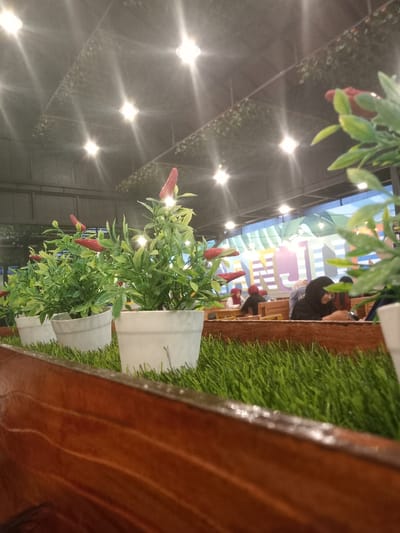 This image captures an indoor dining or cafe environment located in Nganjuk, Indonesia. The foreground features a polished wooden planter box containing artificial green turf, on which a row of small artificial plants in white pots is arranged. Each plant displays green foliage and red, chili-shaped ornamental fruits. Beyond this, in the midground, multiple individuals are seated at tables. One woman on the right, wearing a black hijab, is observed looking downwards. Other patrons are visible further back at light-colored wooden tables. The background wall exhibits a large, colorful mural with abstract geometric patterns in light blue, green, pink, and white tones. The ceiling, dark in color, is decorated with an artificial green leafy net and numerous bright, star-flared circular overhead lights. Horizontal string lights are also present in the background, suspended above the dining area. The scene is illuminated by these artificial lights, creating a bright indoor ambiance.