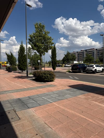 The image shows a street scene in Getafe, Spain, on a sunny day with scattered white clouds in a blue sky. The perspective is from street level, looking down a sidewalk paved with pinkish-brown tiles and grey ribbed strips, possibly indicating a pedestrian walkway with tactile paving.
Several cars are parked along the street, including a white SUV and a dark-colored SUV. In the background, modern buildings with visible windows and balconies rise up. Lush green trees and shrubs are planted along the sidewalk and street, providing shade. Streetlights are visible, indicating an urban environment.
The time of day appears to be midday or early afternoon, given the strong shadows cast by the trees and buildings. The weather is pleasant and sunny. There is no visible text in the image, except for license plates on the cars, which are not legible. The scene is peaceful and ordinary, depicting a typical day in a city.