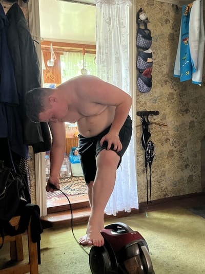 A young, shirtless man with dark hair is vacuuming a room. He is wearing black shorts and is barefoot. The room appears to be in a state of disarray, with clothes hanging on a rack and a patterned wallpaper. A vacuum cleaner, red and black, is on the floor. There are also bottles of water and a trash bag visible in the background. The lighting suggests it is daytime, with sunlight streaming in from a window. The scene appears to be indoors, likely in a residential setting in Sad. Uchastki, Russia.