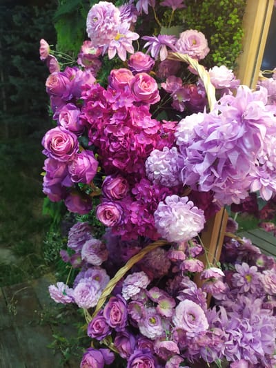 A close-up, high-angle shot features a lush display of artificial flowers in various shades of purple and pink. The flowers are arranged in a cascading fashion, spilling out of woven baskets and against a backdrop of dark green foliage. The textures vary from the tightly packed petals of hydrangeas and roses to the more open, layered petals of chrysanthemums and dahlias. The lighting appears to be ambient, creating soft shadows and highlighting the different colors and forms of the blooms. The overall impression is one of abundance and decorative artistry. The image does not provide any specific identifiable landmarks or features that would definitively place it within the city of Sosnovka, Kyrgyzstan.