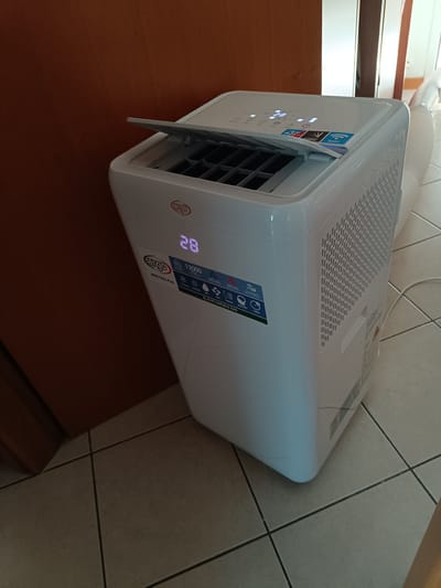 The image shows a white portable air conditioner unit with an open lid, displaying a digital temperature of 28 degrees. It is positioned on a tiled floor indoors, next to a wooden doorframe. Stickers on the unit indicate features such as "ARGO KALL PLUS", "13000 BTU/H", "COOLING", "HEATING", "DRY", "FAN", "SWING", "MEMORY", and "TIMER". The unit also has a Wi-Fi symbol and an energy efficiency rating. The overall environment appears to be a domestic interior. There is no indication of the specific location being Sant'Andrea in Besanigo, Italy, or any people or actions occurring in the scene.