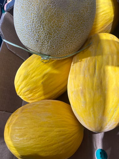 The image shows a close-up, overhead view of several cantaloupe melons stacked in a cardboard box, likely at a market stall in Bel Air, Spain. The melons vary in size and stage of ripeness. One cantaloupe at the top has a distinct netting pattern and a light green vine attached. Below it, three other smooth-skinned yellow melons are visible, one to the left and two to the right, partially obscuring each other. The lighting suggests it is daytime, with bright sunlight casting shadows. The background consists of the cardboard box and a hint of other items in the market. There are no people or animals present in the image.