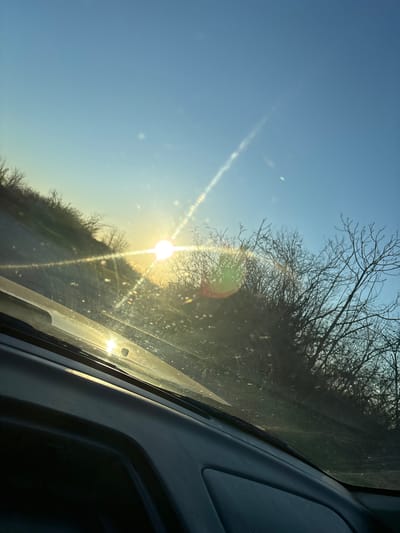 The image is taken from inside a vehicle, looking out through the windshield. The sun is low in the sky, creating a strong glare and lens flare. Part of the dashboard and window frame are visible on the right side of the image. Outside the windshield, there's a view of trees and sky. The sky is a clear, light blue color. The trees are bare, without leaves, suggesting it might be winter or early spring. The scene appears to be rural or on the outskirts of a town, as there are no buildings visible. The location is reported as Suhache, Bulgaria, but there is no visual information to confirm that.