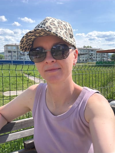 A medium shot captures a woman wearing a leopard-print baseball cap and sunglasses, seated outdoors. She is looking directly at the camera. Her head is slightly tilted, and she has a neutral expression. She is wearing a light purple ribbed tank top. The background shows a green metal fence, grass, and several multi-story buildings in the distance. The sky above is blue with scattered white clouds. The scene appears to be in a residential area, possibly in the city of Kuldur, Russia.