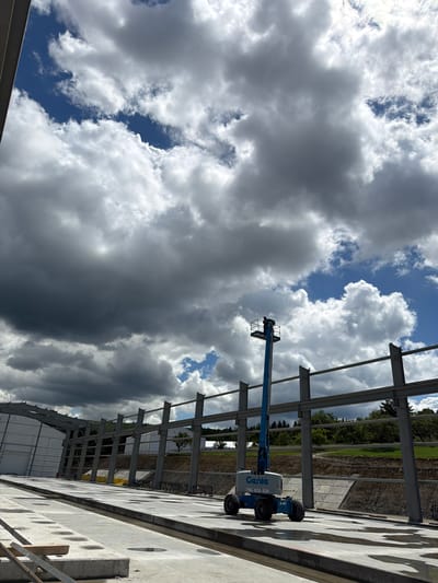 In Janowice, Poland, a construction site is captured under a dramatic sky filled with dark, heavy clouds interspersed with patches of blue. A blue Genie aerial work platform, with the model number S-85 visible, is positioned on a wet concrete surface. The platform is extended, and a person can be seen in the basket at the top. In the background, a large steel-framed structure is under construction, with rows of structural beams and columns visible. The ground is a mix of concrete, exposed soil, and gravel. Green trees and rolling hills are visible beyond the construction area.