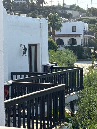 The image depicts an outdoor scene in a Mediterranean setting, likely Chersonisos, Greece, based on the provided location context. The primary subjects are buildings with white stucco exteriors, featuring architectural elements such as arched windows and terracotta-tiled roofs. A dark wooden staircase with a modern design is prominently featured in the foreground, leading upwards and obscuring a portion of the white buildings. Lush green foliage and trees, including palm trees, are visible throughout the scene, suggesting a warm climate.

In the middle ground, another building with distinct arched windows and balconies is visible. Beyond this, more buildings and trees stretch into the background, with some suggesting a hilly or tiered landscape. Power lines are visible in the upper left of the image, indicating the presence of modern infrastructure.

The lighting suggests it is daytime, with sunlight casting shadows and highlighting textures on the buildings and foliage. The overall mood is tranquil and serene, characteristic of a holiday or residential area in a sunny climate. No people are clearly visible, though there are hints of colorful fabric on the stairs which might suggest discarded items or part of a resting place. There is no visible text in the image. The scene appears to be a view from a balcony or higher level, looking down onto a street or courtyard area.