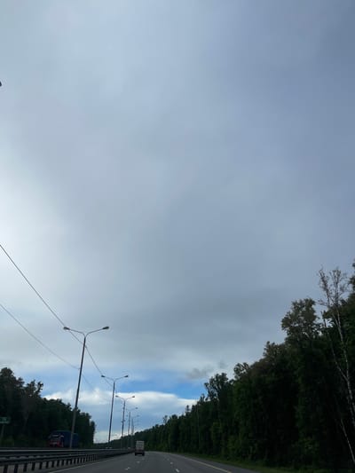 The image shows a view from a highway in or near Бавыкино, Russia. The sky is predominantly overcast, with some patches of blue visible through breaks in the clouds. A line of street lights runs along the right side of the highway. The road itself appears to be multi-lane. A truck and a car are visible in the distance. The landscape is bordered by trees on both sides.