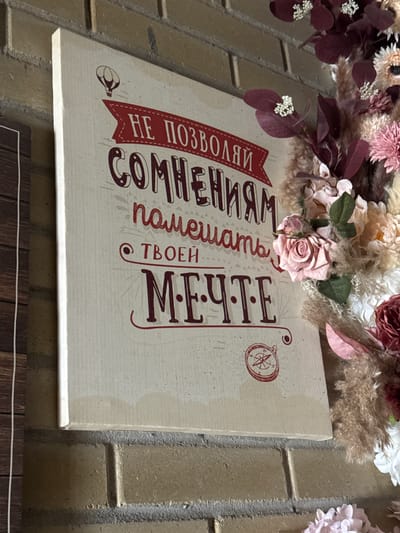 The image shows a decorative sign hanging on a brick wall, partially obscured by floral arrangements at the top. The sign is cream-colored with text in Russian. There's a banner at the beginning of the text. There is a compass drawing and what seems to be a hot air balloon sketch. The surrounding decor includes roses and other flowers. The photo may have been taken in the city of Zamankul, Russia, although this detail doesn't affect the description.