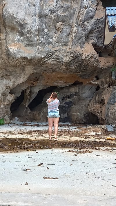 The image depicts a woman standing at the entrance of a cave in Thap Put, Thailand. The cave is formed in a large rock formation, with multiple openings visible. The woman is wearing denim shorts and a striped shirt, and appears to be looking into the cave. The foreground consists of a concrete surface with scattered debris. A building or structure is visible atop the rock formation to the left of the cave entrance.