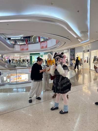 Inside a bright, modern shopping mall, a group is captured, primarily showcasing a person in a striking costume. The individual is adorned with a black dress, white tights, and light blue leg warmers paired with black shoes, accompanied by an elaborate headpiece with a white furry collar. Nearby, another person in a black shirt and loose beige pants stands. The backdrop is a multi-level mall with various stores, including "KEYLAI" visible in the upper level.