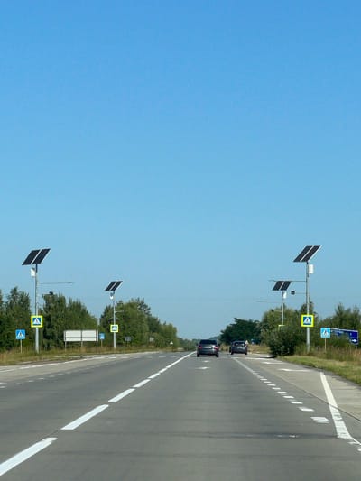 This is a photograph taken from the perspective of someone in a vehicle on a paved road with clear lane markings. The sky is a solid, bright blue with no clouds. The road appears to be a highway or a major road with trees lining both sides.
On the left side of the road, there are two pedestrian crossing signs with blue backgrounds and white pedestrian icons. One sign is higher on a pole with a solar panel above it. Next to these is a larger, blank white rectangular sign. Further down the road, another solar-powered pole with a pedestrian crossing sign is visible.
On the right side of the road, there are also solar-powered poles with pedestrian crossing signs. One of these poles also has a blue road sign with a white pedestrian icon and text that appears to say "И ГОРОД" (which translates to "To City") with an arrow pointing to the right and a number "1".
There are two dark-colored cars driving away from the camera on the right lane of the road. The environment suggests a rural or semi-rural setting with dense green trees visible on both sides of the road. The scene is well-lit, indicating it's daytime. The location is identified as Mala Zubivshchyna, Ukraine.
