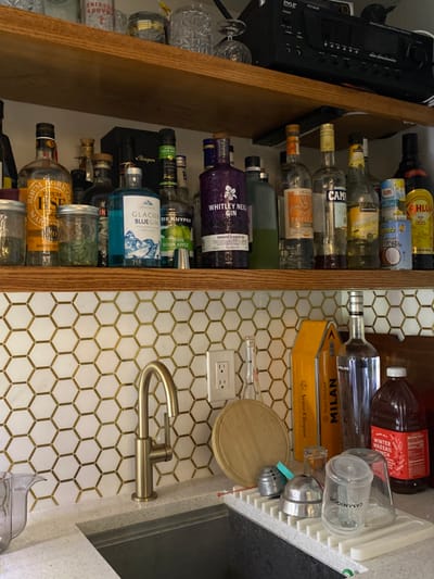The image depicts an indoor residential setting in Ridgefield, United States, specifically a home bar or kitchen area. The scene is dominated by two light-colored wooden shelves mounted above a countertop.
The lower wooden shelf displays approximately twenty bottles of various alcoholic beverages, including clear, colored, and dark spirits, alongside several glass jars. Identifiable brands include Rhum Agricole HSE, Glacier Blue Gin, De Kuyper Sour Apple Pucker, Whitley Neill Rhubarb & Ginger Gin, Campari, Kahlua, and Kirkland Signature Vodka. Two cans, one labeled "Dole" and the other "Goya Coconut Milk," are also present. The upper shelf holds miscellaneous clear glassware and a black electronic audio receiver.
Below the shelves, a backsplash features white hexagonal tiles with gold-colored grout lines. An electrical outlet with two receptacles is centered on this tiled surface. A light-colored countertop with an integrated dark-colored sink basin and a modern brushed gold or brass-toned gooseneck faucet occupies the foreground. To the right of the sink, on the countertop, a small round wooden cutting board, an empty clear glass bottle, and a tall yellow box labeled "Veuve Clicquot" and "MILAN" are arranged. A large plastic bottle containing a red liquid, labeled "WINTER WASSAIL PUNCH," stands next to these items. A white plastic dish drying rack holds washed barware, including metal shakers or strainers and two clear glass tumblers, one marked "CASAMIGOS." A clear plastic pitcher is partially visible on the far left of the countertop. No individuals are present, and no actions or interactions are depicted.