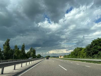 The media file depicts a view from a car driving on a highway, likely in Mytnytsia, Ukraine. The scene is dominated by a dramatic sky filled with dark, heavy storm clouds interspersed with patches of brighter, broken clouds and a few glimpses of blue sky. This suggests impending rain or a recent shower.

The highway itself is paved and has multiple lanes, separated by white dashed lines. A silver metal guardrail runs along the left side of the road, separating the lanes from the grassy shoulder and a line of trees. On the right side of the highway, the shoulder is also bordered by lush green trees and vegetation, with a few more tall trees visible further in the distance.

Several vehicles are visible on the highway, including a dark-colored car in the lane ahead, and a white car further in the distance on the left. A yellow bus can be seen in the far distance on the left. Streetlights are positioned periodically along the right side of the highway.

A small, light-colored building with a blue roof is situated on the right side of the highway in the mid-ground, possibly a rest stop or service area. Blue road signs with white symbols are also visible along the road. The time of day appears to be daytime, given the overall brightness, but the stormy clouds cast a somewhat dim light.

The overall impression is one of a journey through varied weather conditions, with the dramatic sky being a prominent feature of the landscape.