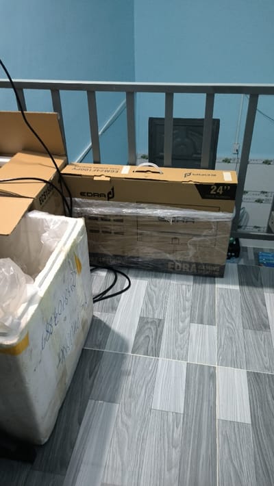 The image shows an interior scene with a light blue wall and a metal railing. The floor is tiled with a wood-grain pattern. A cardboard box labeled "EDRA Gaming Monitor 24" wide screen" is laying on the floor. A white styrofoam box with writing on it is also on the floor. A black electrical cord runs across the floor. There is a black television behind the railing, but only the back is visible. The image is likely taken in a residential building in Thành phố Hồ Chí Minh, Vietnam. The scene is likely a living room or bedroom.