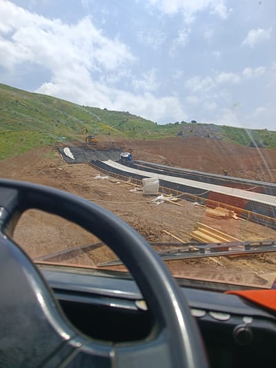 The image shows a construction site on a green, hilly landscape under a cloudy sky. In the foreground, the black steering wheel of a vehicle is visible, suggesting the photo is taken from the driver's perspective. The middle ground reveals ongoing construction work with heavy machinery, including an excavator. There are mounds of dirt, building materials like stacked wood, and what appears to be some sort of track or road being constructed, possibly with black matting or panels. A few workers in yellow vests are present on the site. The background features a lush green hillside dotted with yellow wildflowers. The location is identified as Nurekata, Uzbekistan.